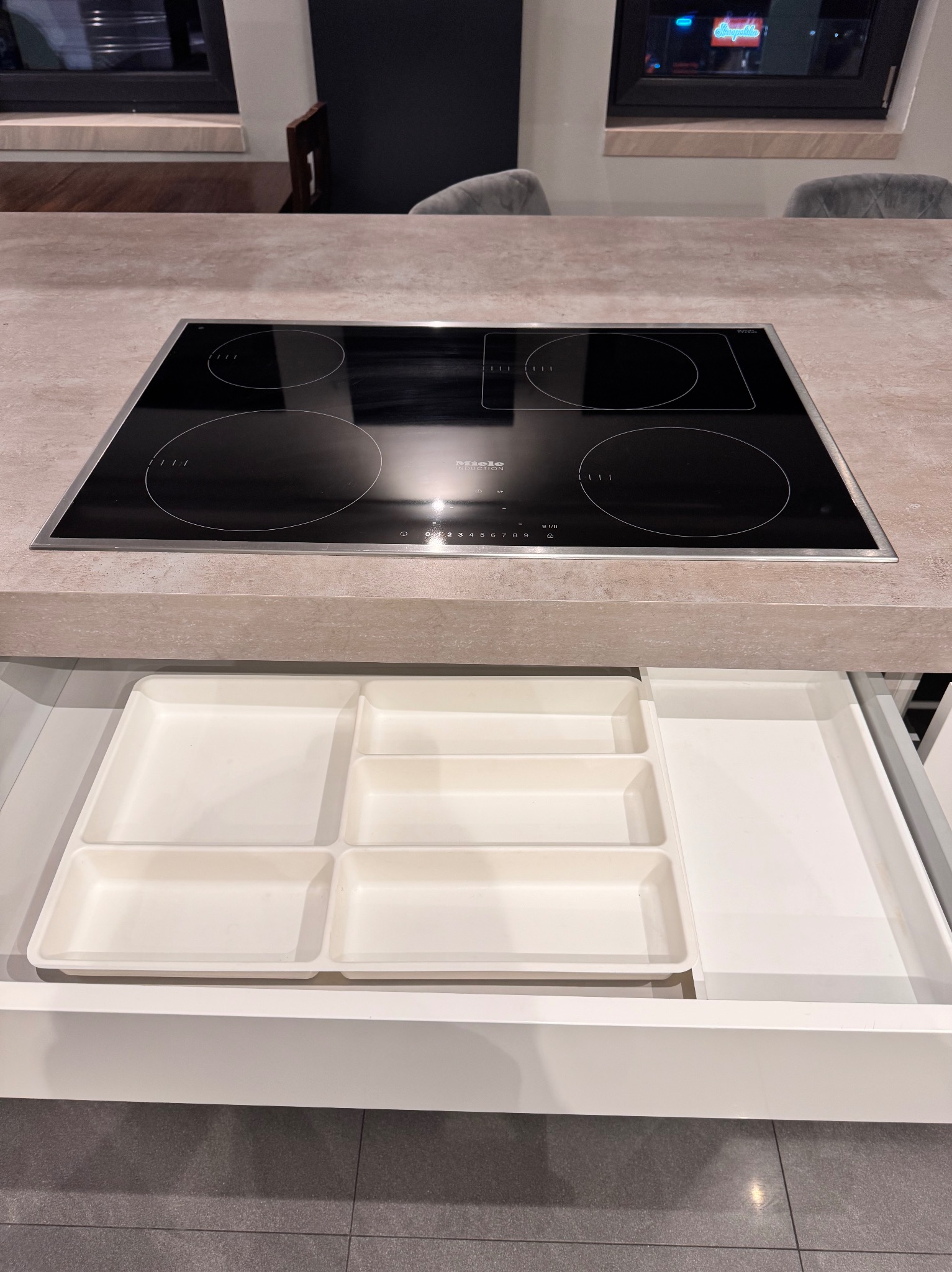 Nowoczesna kuchnia z blatem z szarego kamienia, czarną płytą indukcyjną Miele i otwartą białą szufladą z przegródkami na sztućce i akcesoria kuchenne.