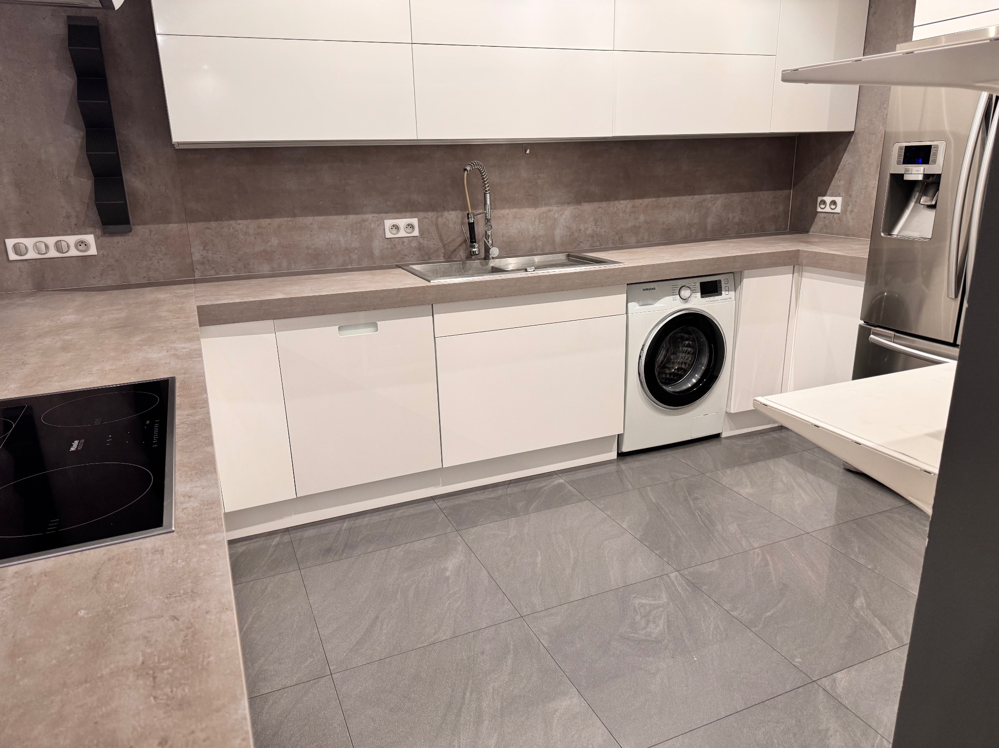 Nowoczesna, minimalistyczna kuchnia z białymi szafkami, szarym blatem i płytkami podłogowymi, z pralką w zabudowie obok zlewu i stalową lodówką.