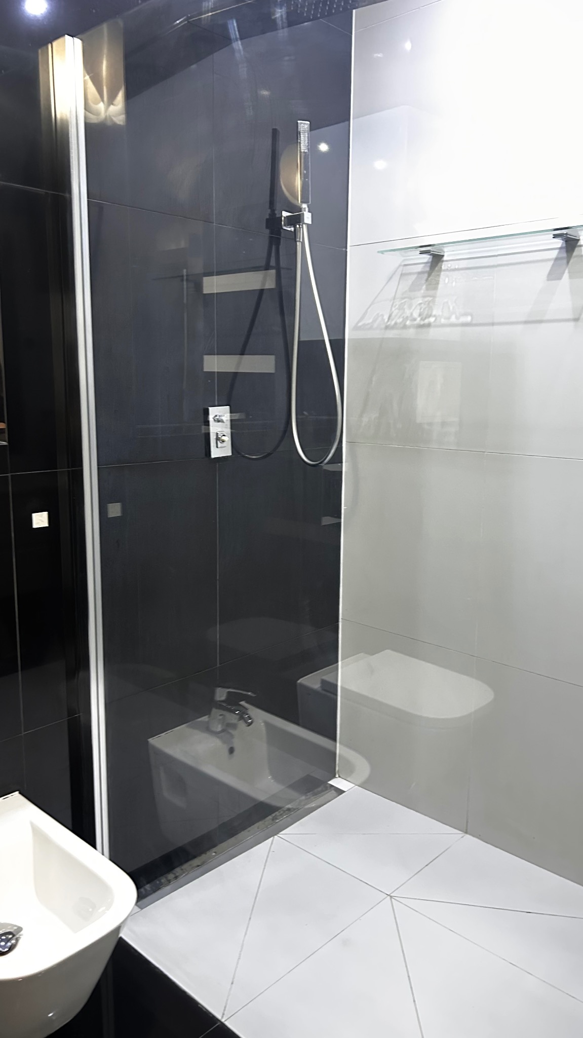 Nowoczesna kabina prysznicowa z czarnymi i białymi płytkami, widoczna armatura i szklane półki, bidet i umywalka w tle, geometryczny wzór płytek podłogowych.