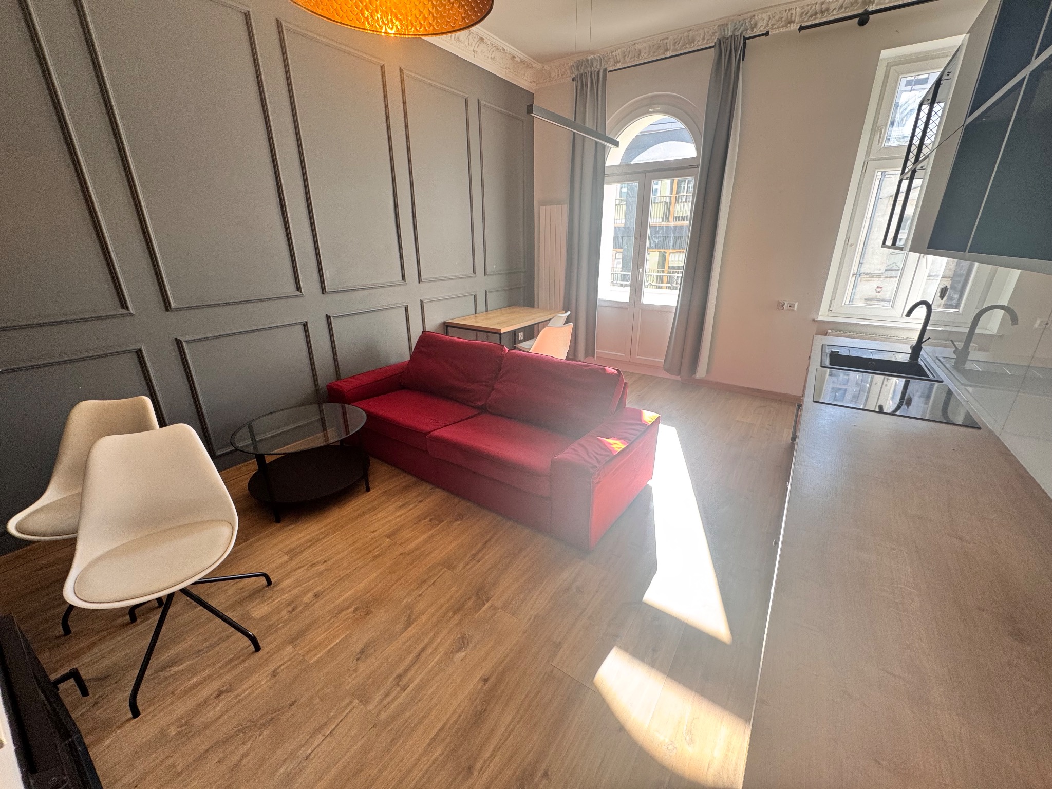 Nowoczesne wnętrze mieszkania z czerwoną sofą, drewnianą podłogą i szarą ścianą z dekoracyjnymi panelami, oświetlone ciepłym światłem lampy i wpadającym przez okno promieniem słońca.