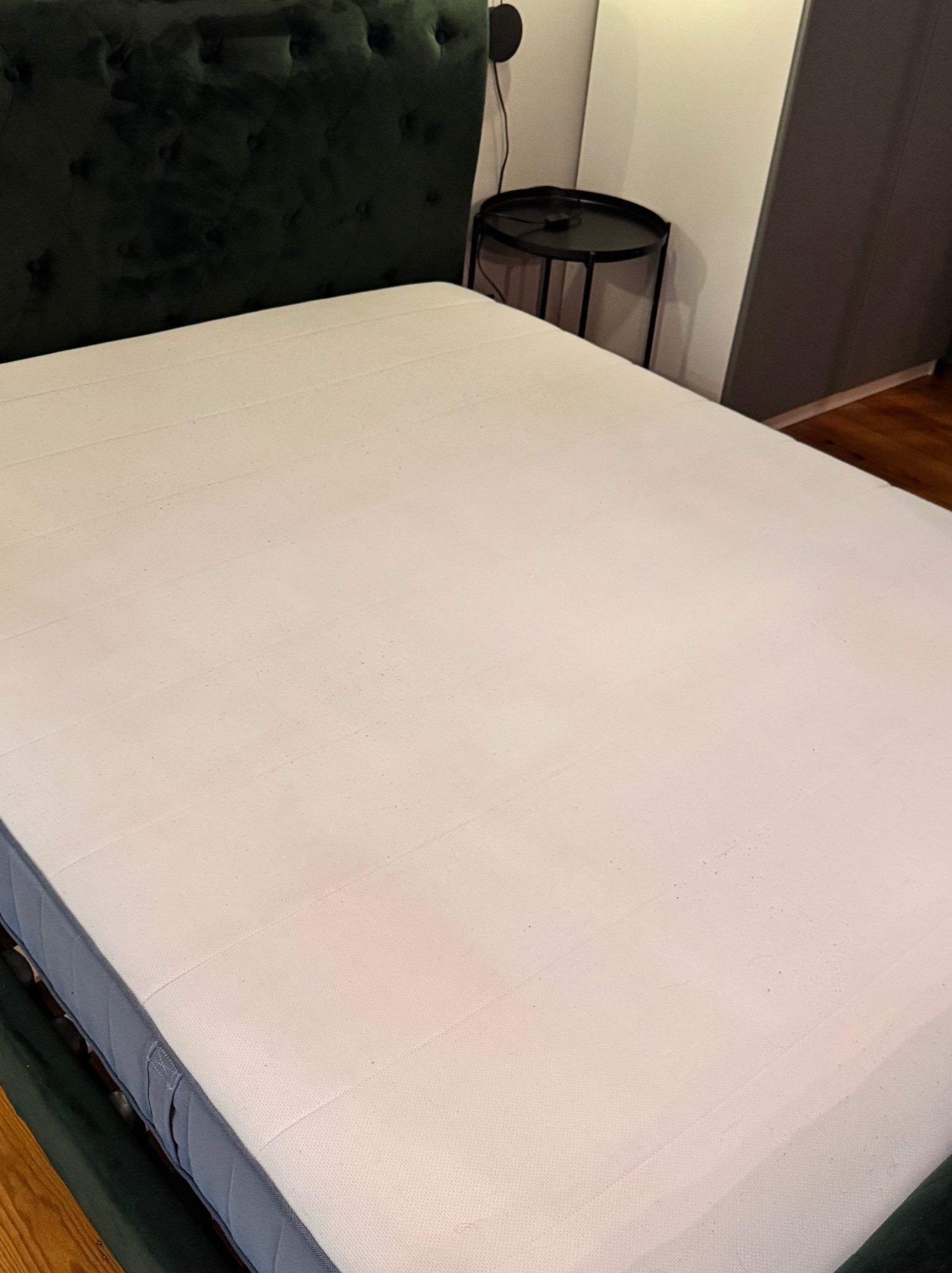 Powierzchnia materaca z widocznymi drobnymi zabrudzeniami, na tle ciemnozielonego wezgłowia i stolika nocnego.