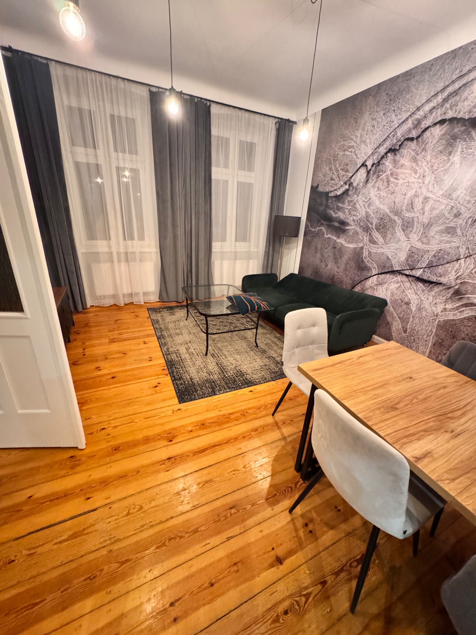 Ustawny salon z drewnianą podłogą, ciemnozieloną sofą, szklanym stolikiem kawowym i drewnianym stołem z krzesłami na tle ściany z abstrakcyjnym wzorem.