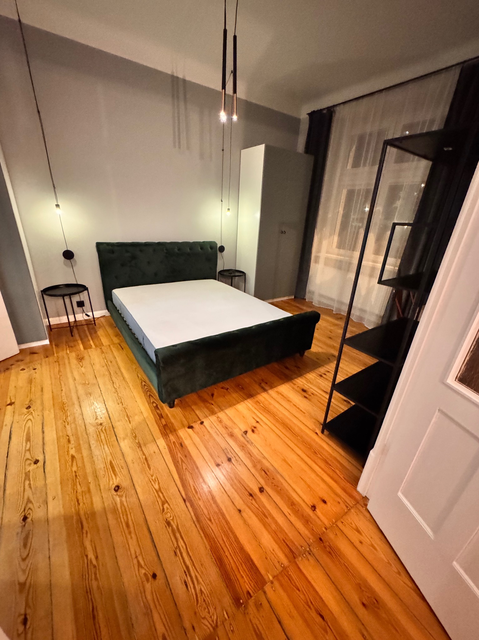 Sypialnia z zielonym, welurowym łóżkiem na drewnianej podłodze, oświetlona wiszącymi lampami i lampkami nocnymi na stolikach.