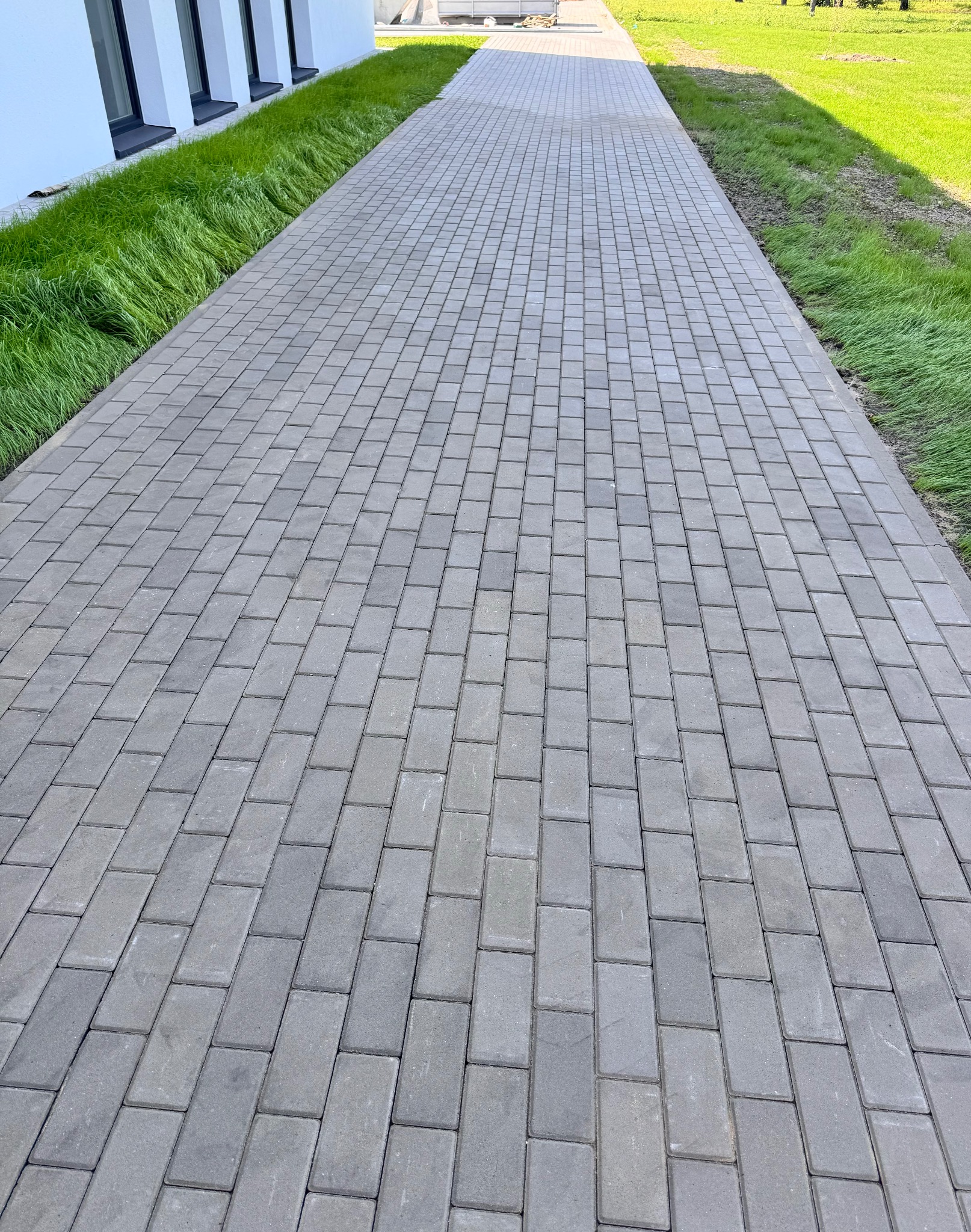 Ułożona, szara kostka brukowa tworząca ścieżkę wzdłuż budynku z zielonym pasem trawy. Widok z perspektywy.