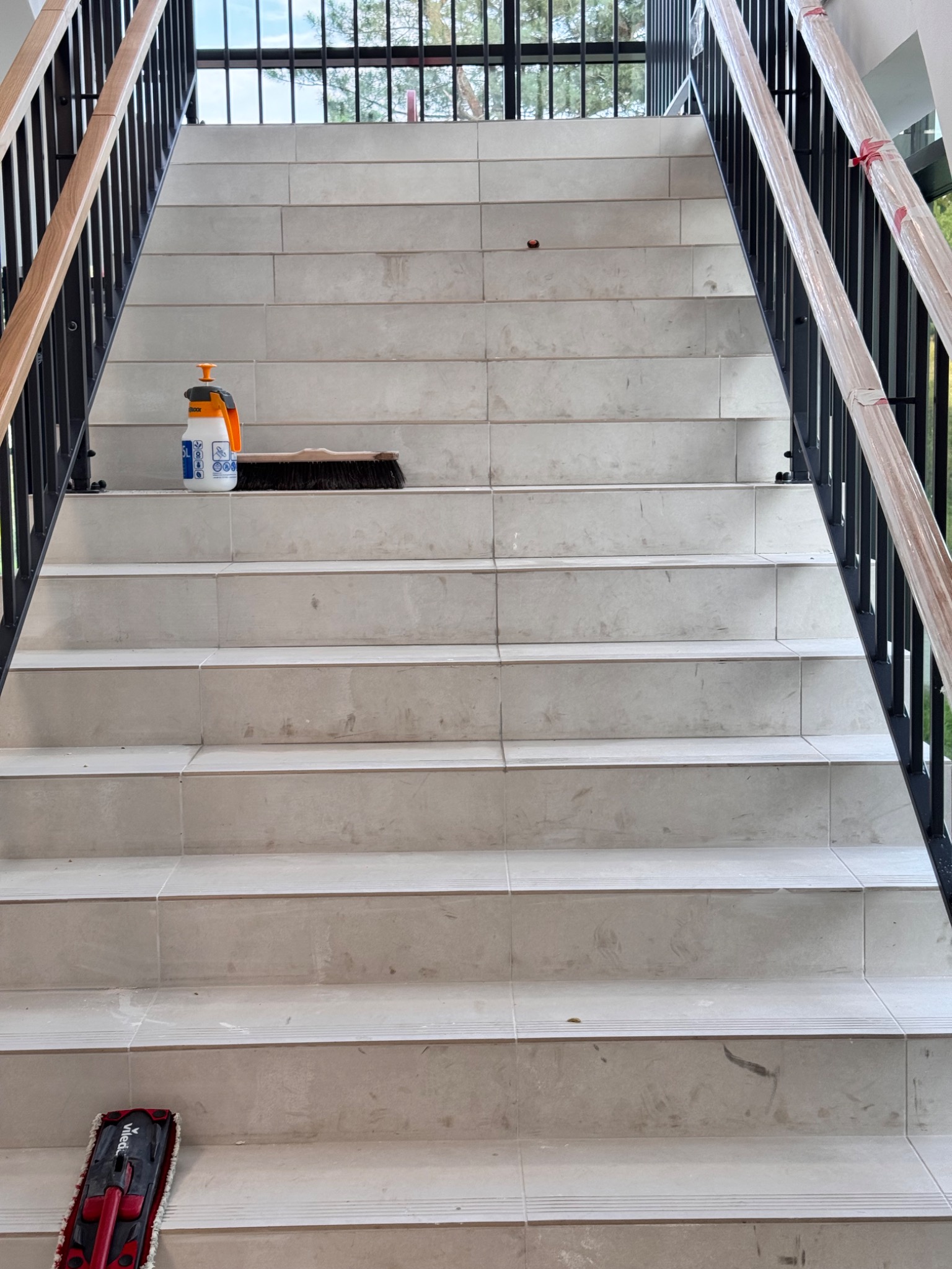 Schody z płytek w trakcie sprzątania: szczotka, mop i butelka ze środkiem czyszczącym. Balustrady z czarnego metalu z drewnianymi poręczami.