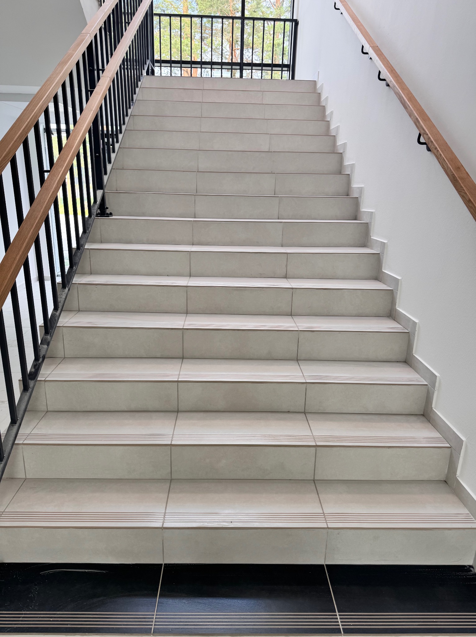 Klatka schodowa z beżowymi płytkami, czarną metalową balustradą i drewnianymi poręczami. Widok z dołu na górę, z widocznym oknem na końcu schodów.