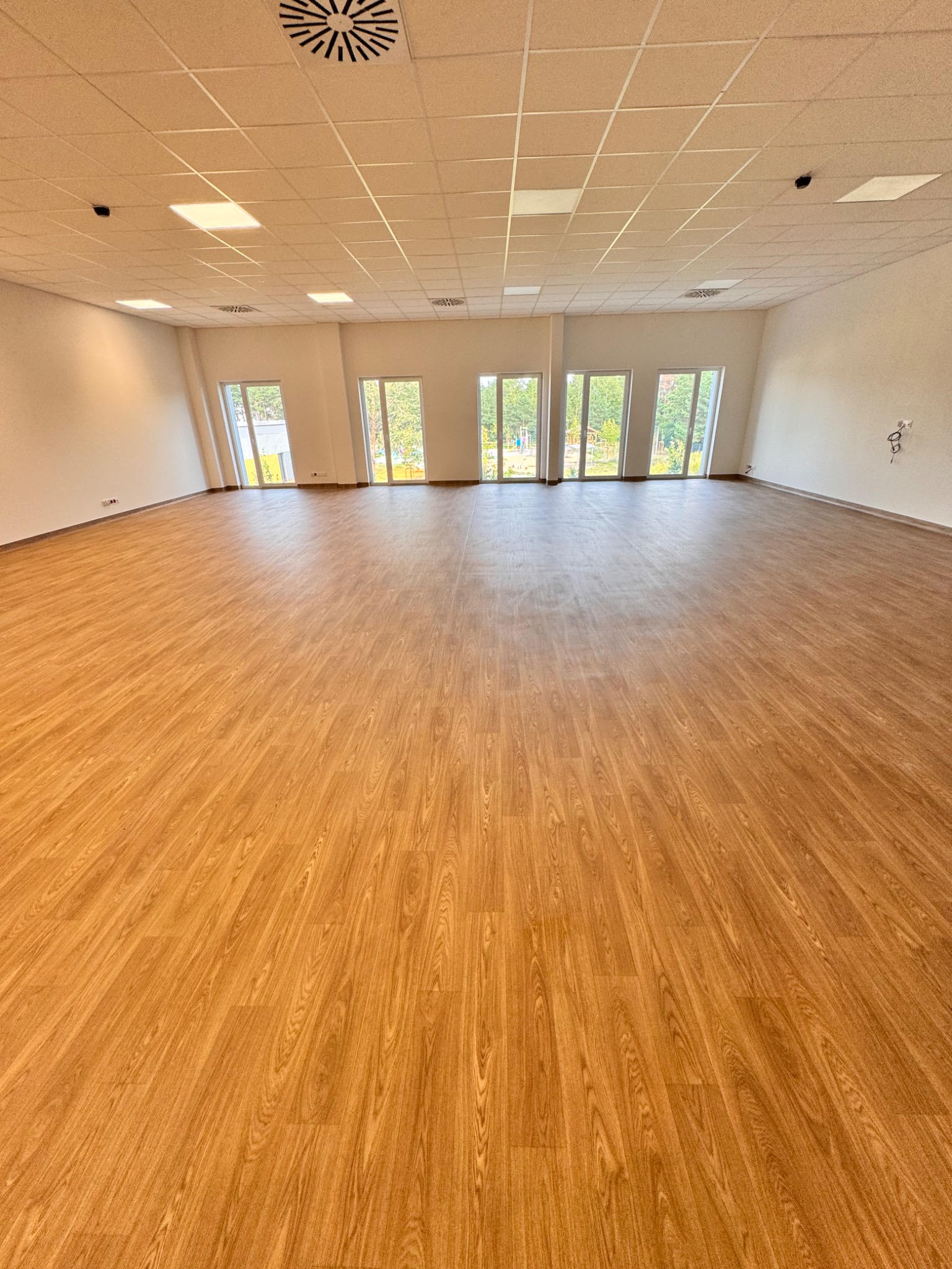 Jasne, przestronne wnętrze po sprzątaniu. Parkiet z wzorem drewna, białe ściany i sufit z oświetleniem. Widoczne okna i elementy wentylacji.