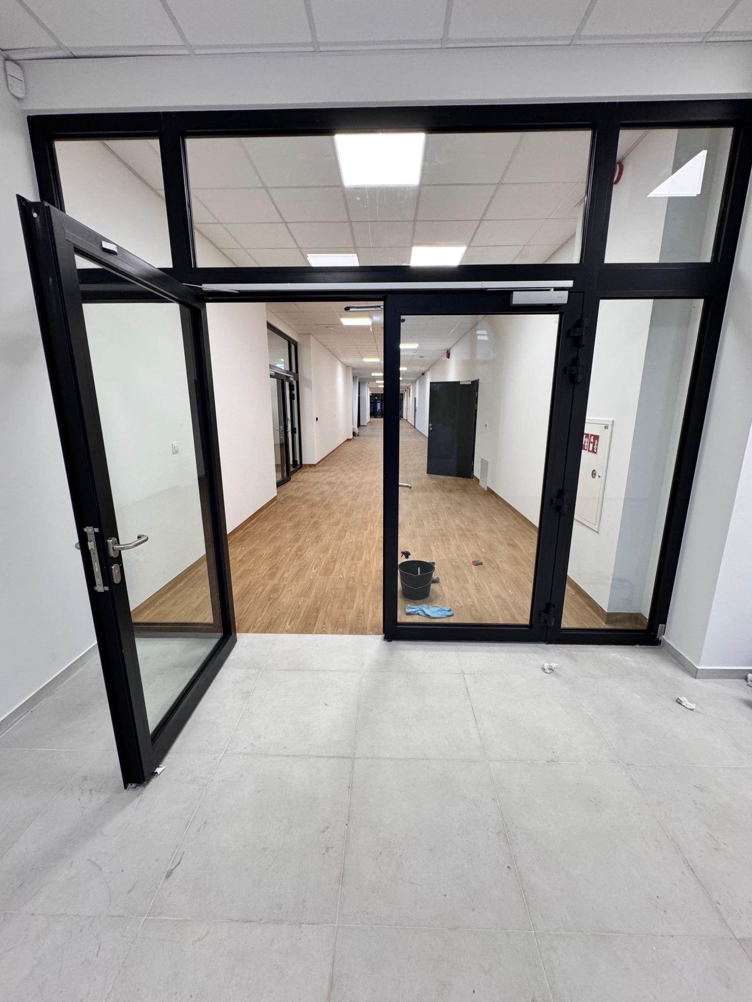 Wnętrze biura po sprzątaniu: otwarte drzwi, widoczny korytarz z czystą podłogą, wiadro i ściereczka w tle, jasne płytki podłogowe. Perspektywa z dołu.