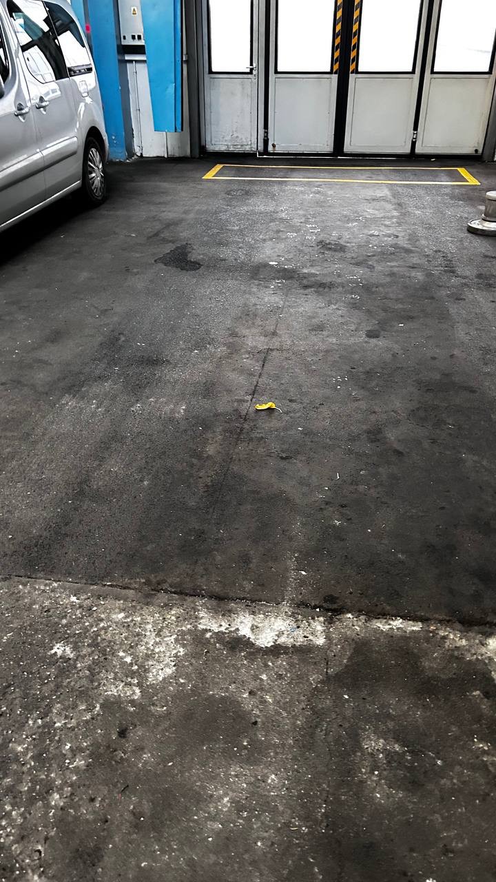 Brudna, betonowa posadzka w hali z żółtą linią, fragmentem samochodu i pojedynczym żółtym liściem. Widoczne plamy i zabrudzenia.