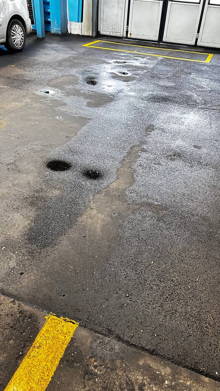 Mokry asfalt z żółtymi liniami i kałużami. Fragment samochodu w lewym górnym rogu. Widoczne ślady po czyszczeniu, sugerujące niedokładne mycie.