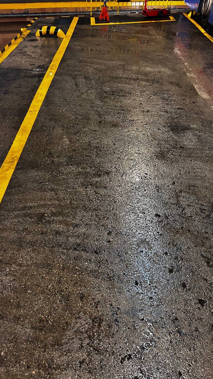 Mokra, ciemna posadzka z żółtymi liniami i odbojnikami. Widoczne ślady po czyszczeniu, wskazujące na świeżo umytą powierzchnię.