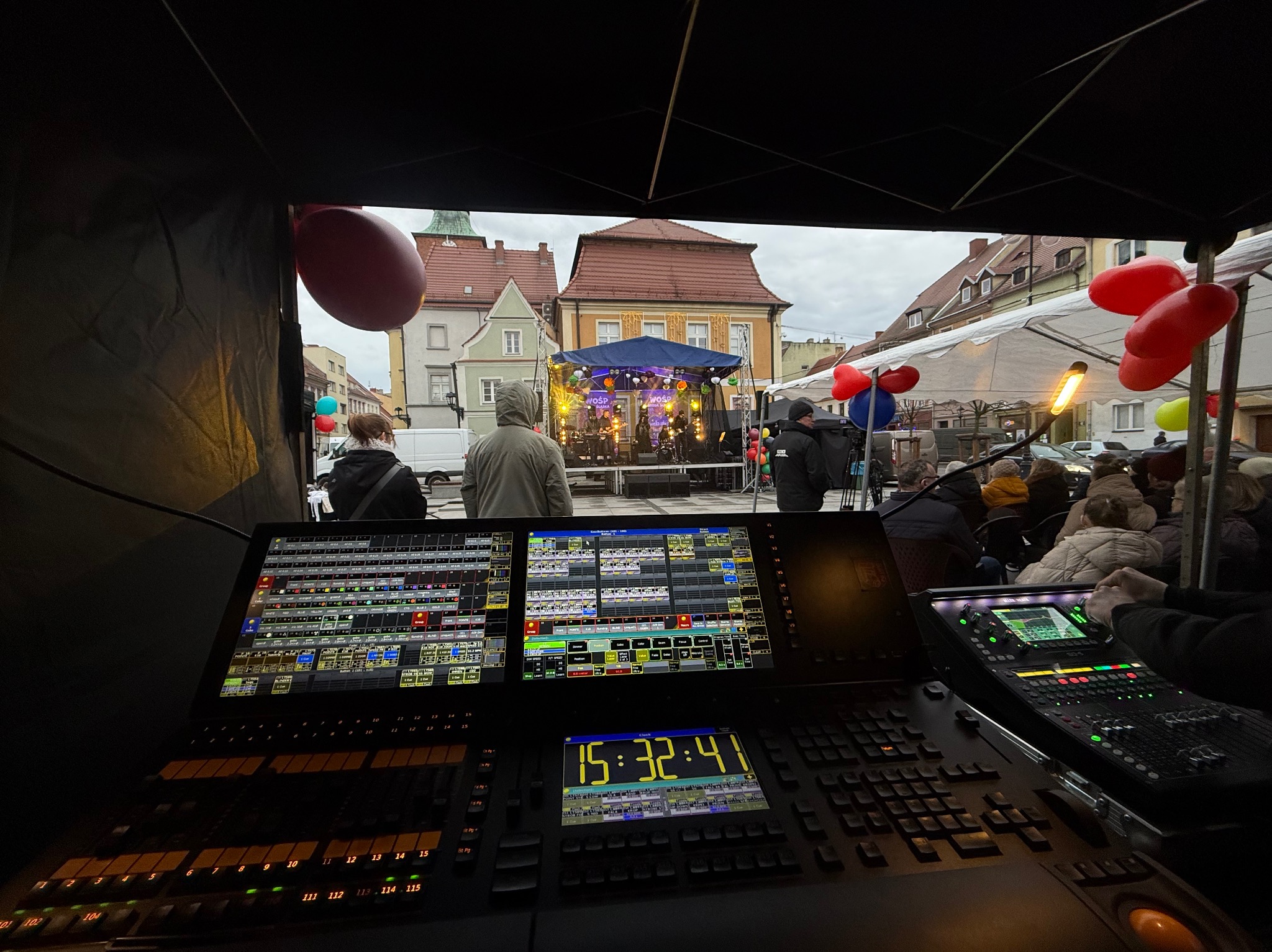 Stanowisko realizatora nagłośnienia i oświetlenia podczas imprezy plenerowej w Szczepanowie, widok na scenę zza konsolety, w tle budynki miejskie i publiczność.