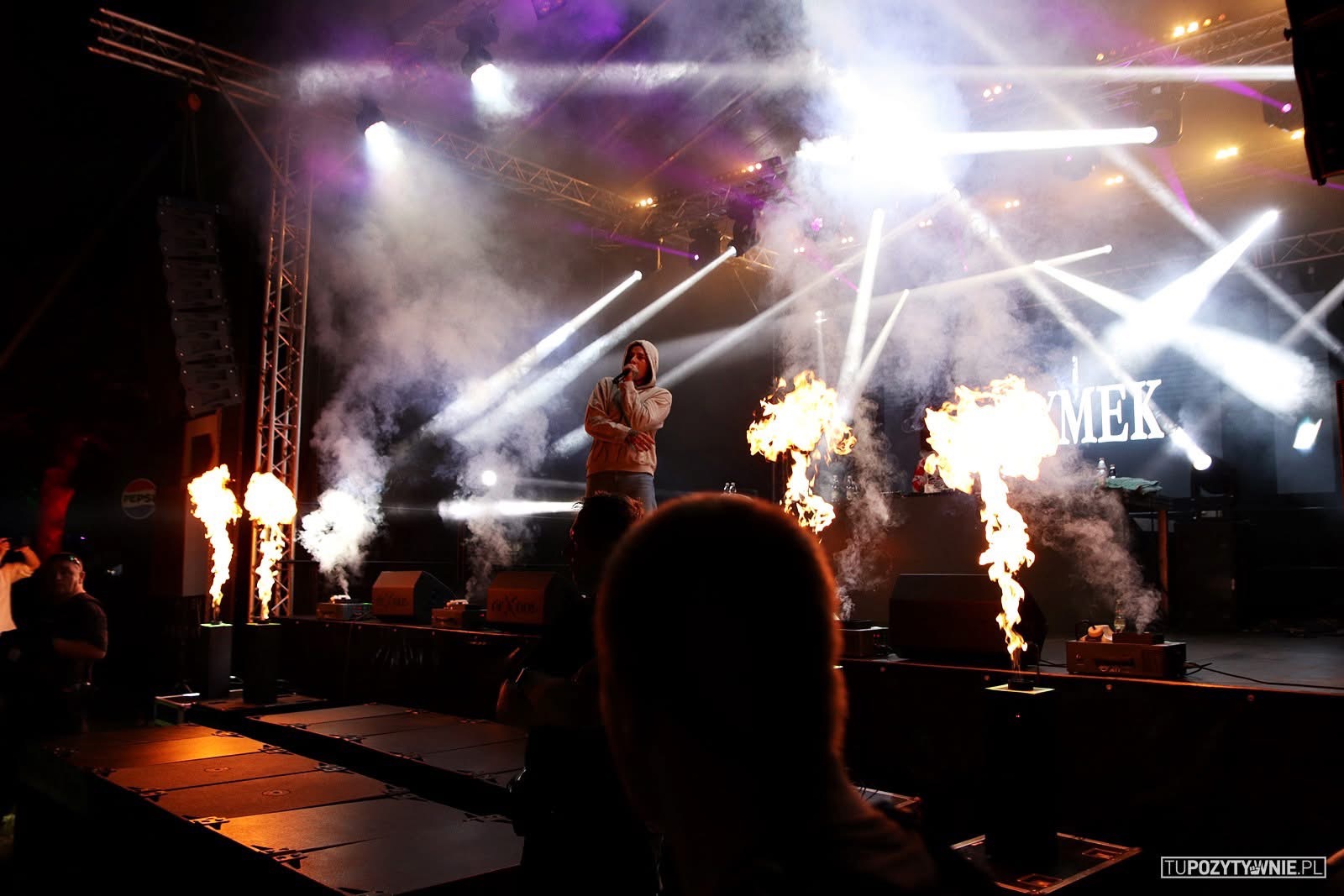 Wokalista w bluzie z kapturem występuje na scenie z efektami pirotechnicznymi i oświetleniem punktowym, widoczna konstrukcja kratownicowa i dym.