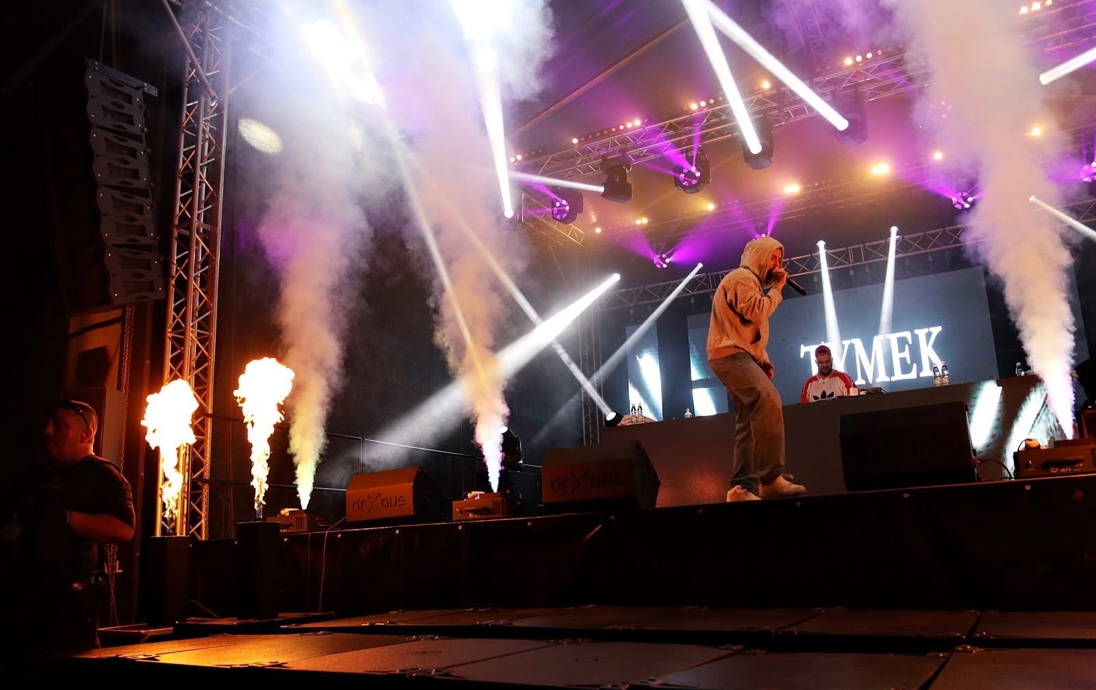 Koncert na scenie: wokalista w bluzie z kapturem śpiewa do mikrofonu, za nim DJ, efekty pirotechniczne i oświetlenie sceniczne tworzą widowiskową atmosferę.