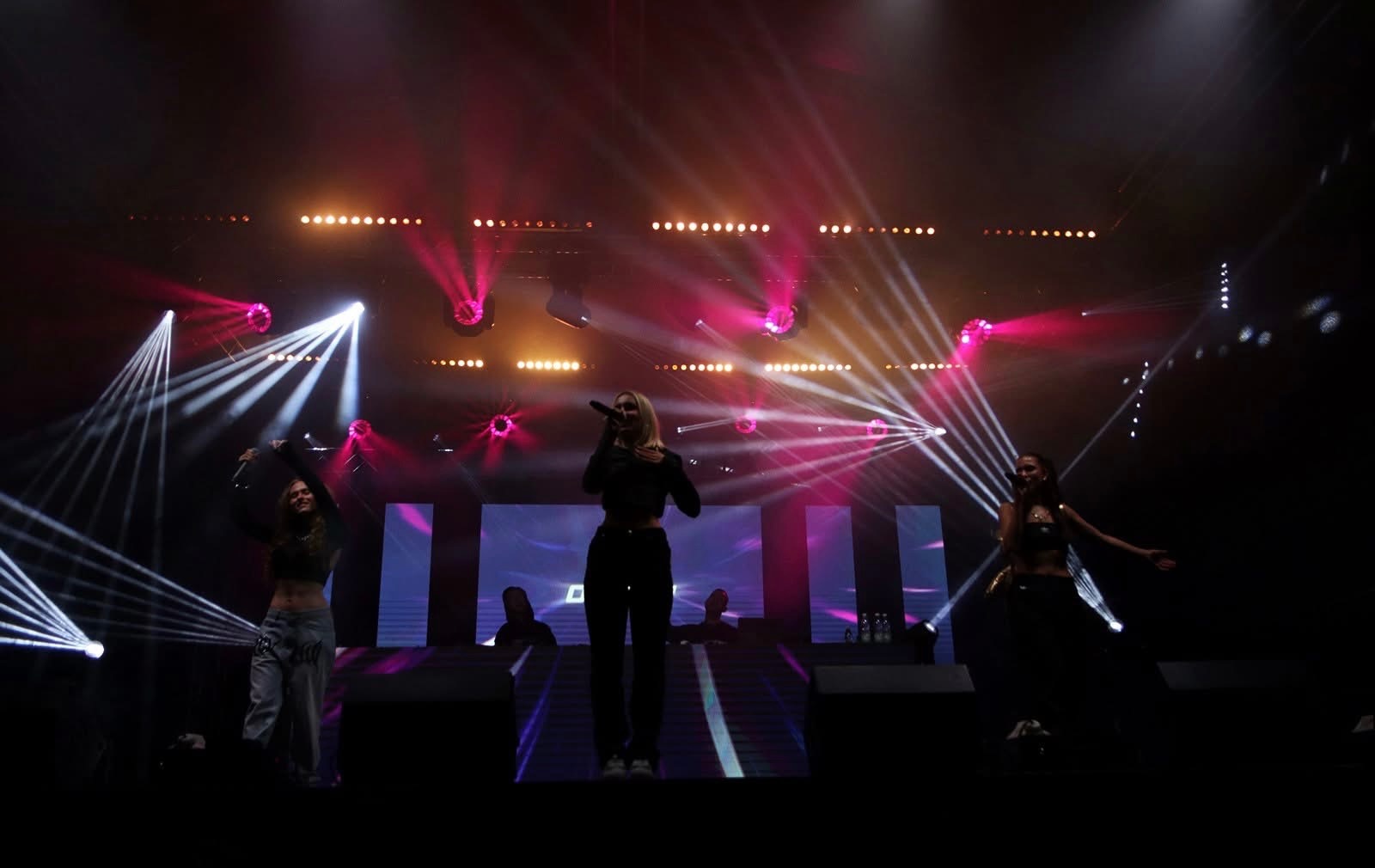 Wokalistka z mikrofonem na scenie podczas koncertu, oświetlona reflektorami i światłami punktowymi, z dwiema tancerkami po bokach.
