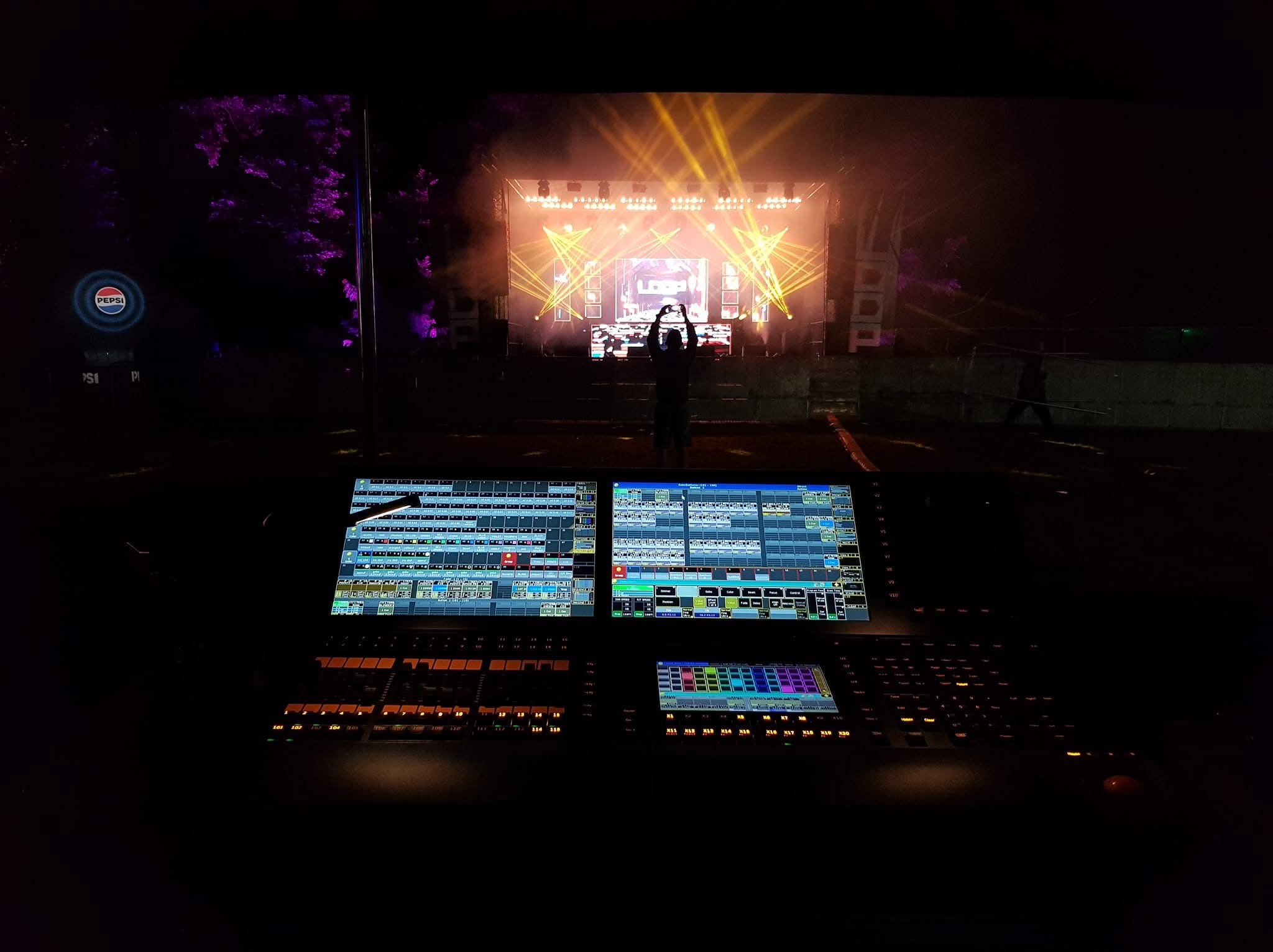 Widok zza konsolety oświetleniowej i dźwiękowej na scenę z efektami świetlnymi podczas koncertu plenerowego, osoba filmuje scenę telefonem.