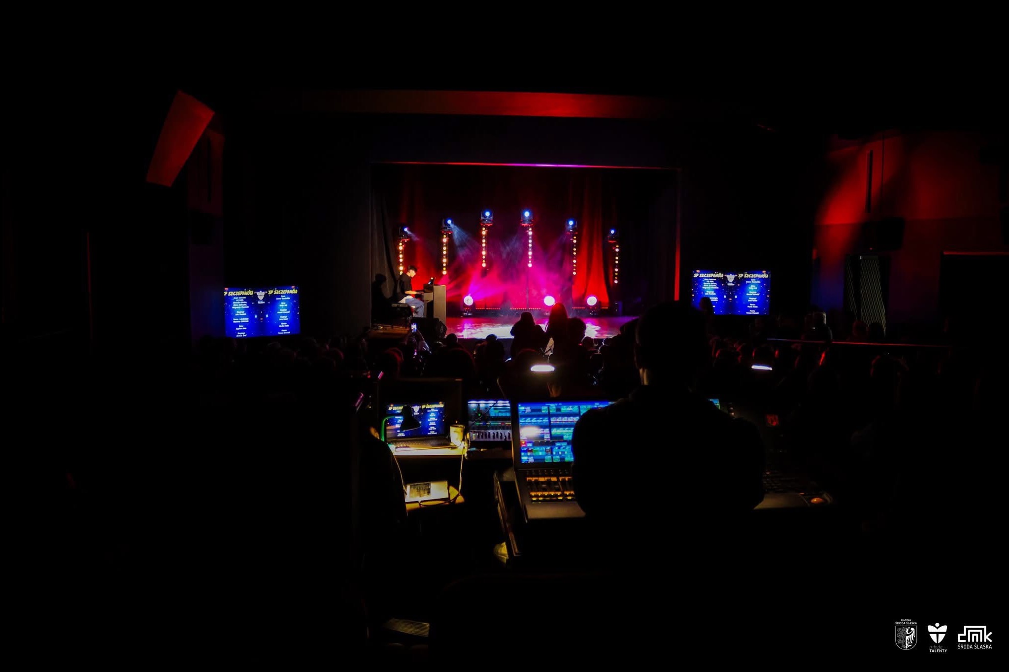 Widok zza kulis na scenę podczas wydarzenia z oświetleniem scenicznym i osobą grającą na pianinie, z widocznymi monitorami i sprzętem kontrolnym.