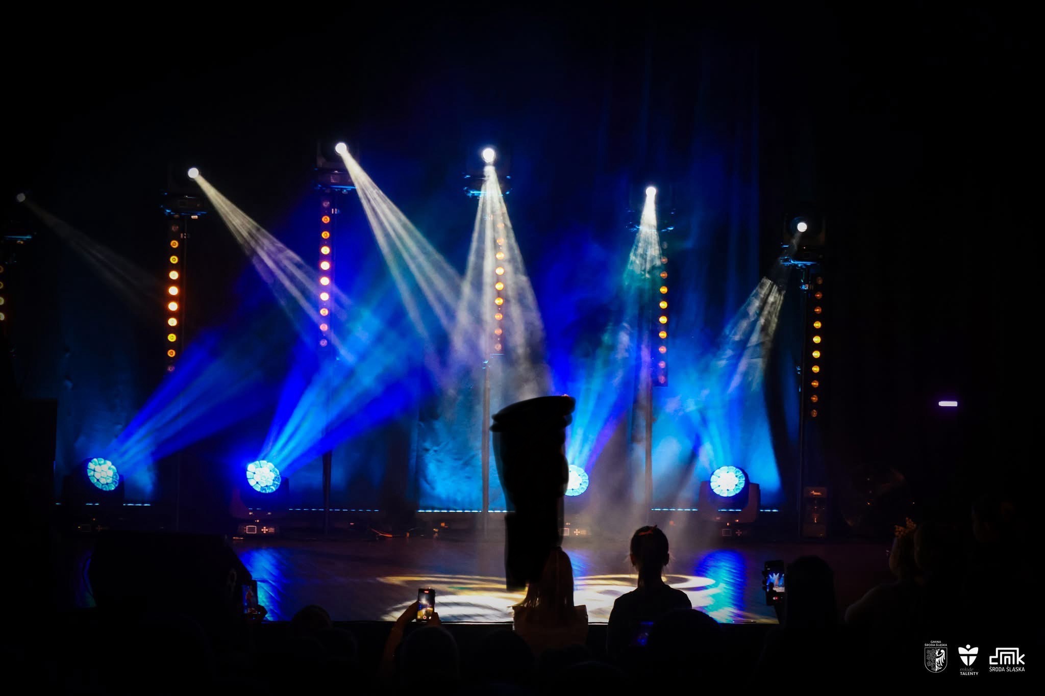 Scena oświetlona niebieskimi i białymi reflektorami podczas wydarzenia, widoczni widzowie w tle.