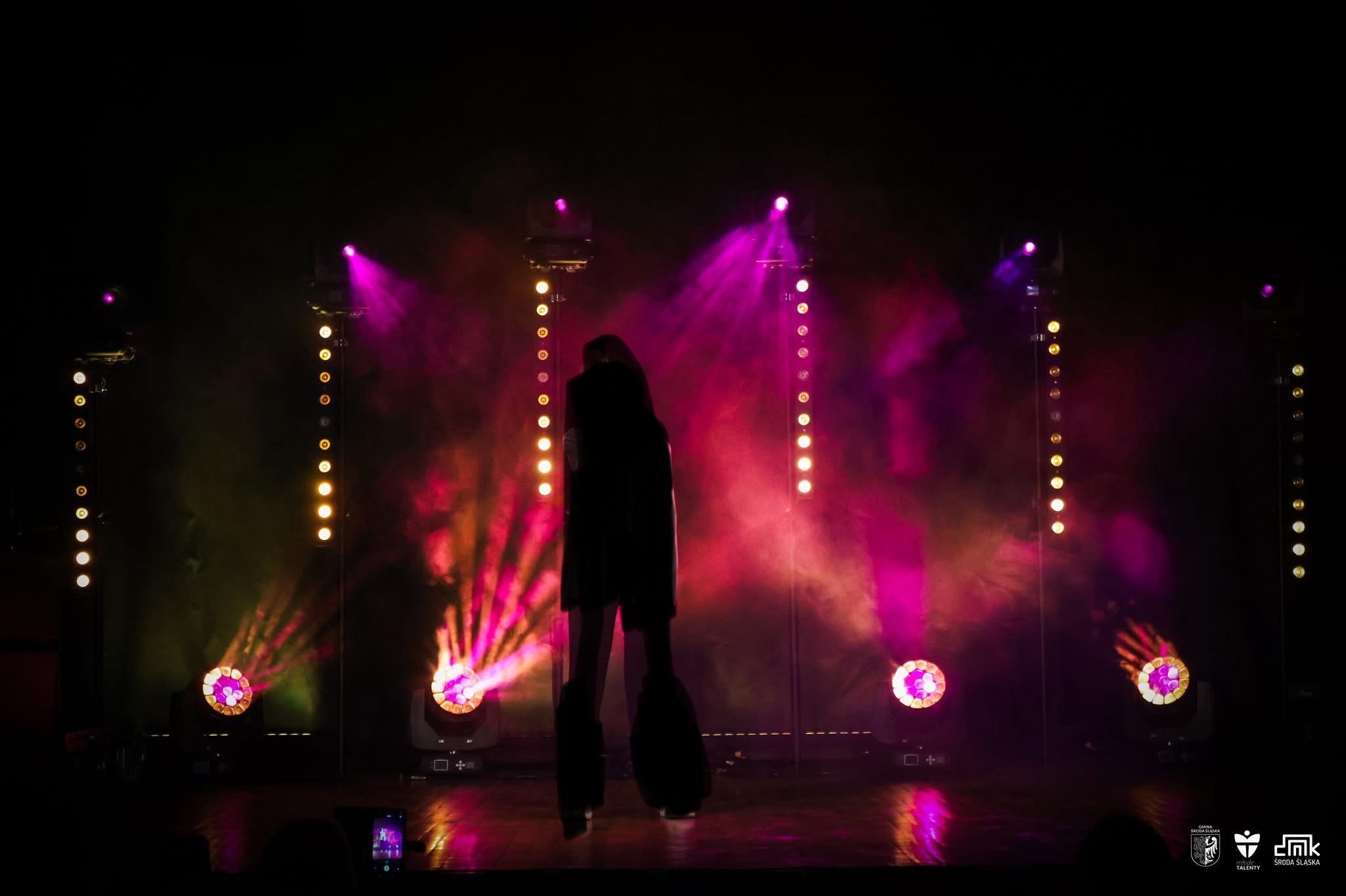 Sylwetka osoby na scenie oświetlonej reflektorami w kolorach różu i pomarańczy, z rzędami małych świateł po bokach.