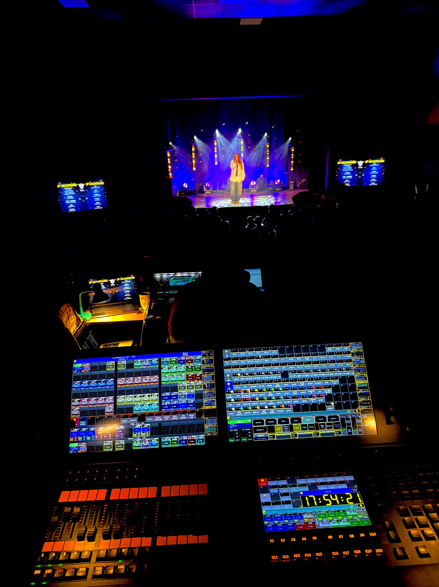 Widok zza konsoli oświetleniowej i nagłośnieniowej na scenę podczas występu artystycznego z oświetleniem punktowym i ekranami bocznymi.
