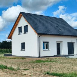 RAFBUD Firma Budowlano-Montażowa - Nowoczesny dom jednorodzinny z białymi ścianami, grafitową dachówką i roletami zewnętrznymi, widok z zewnątrz w słoneczny dzień.