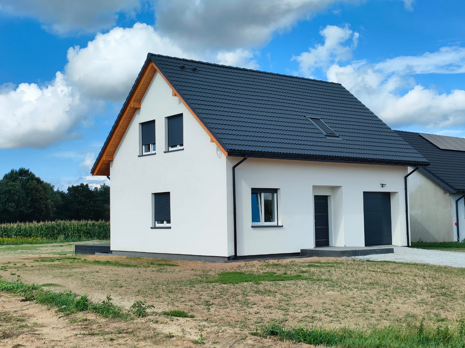 Nowoczesny dom jednorodzinny z białymi ścianami, grafitową dachówką i roletami zewnętrznymi, widok z zewnątrz w słoneczny dzień.