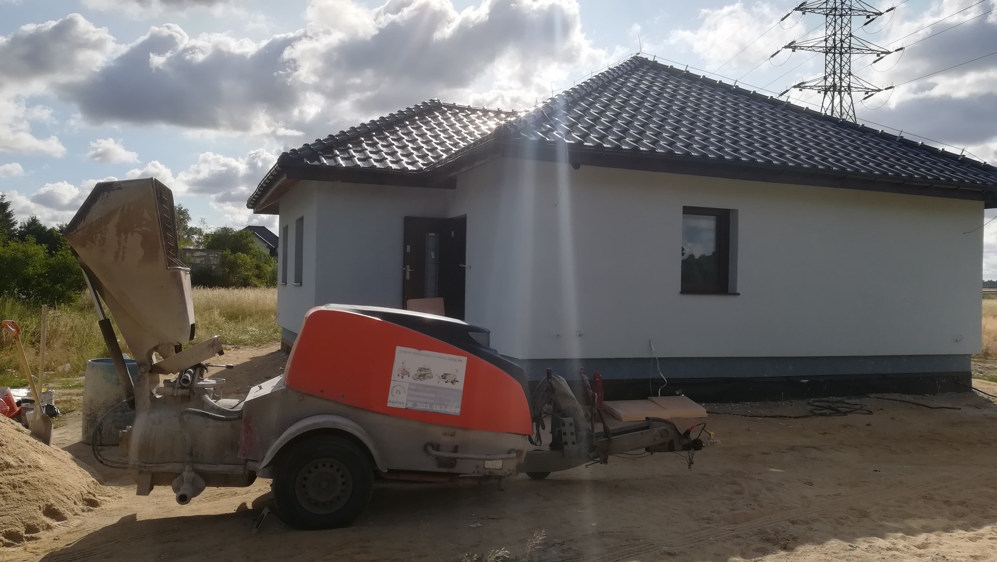 Maszyna do tynkowania elewacji stoi przed nowo wybudowanym, parterowym domem z ciemną dachówką, w tle słupy wysokiego napięcia i pochmurne niebo z promieniami słońca.