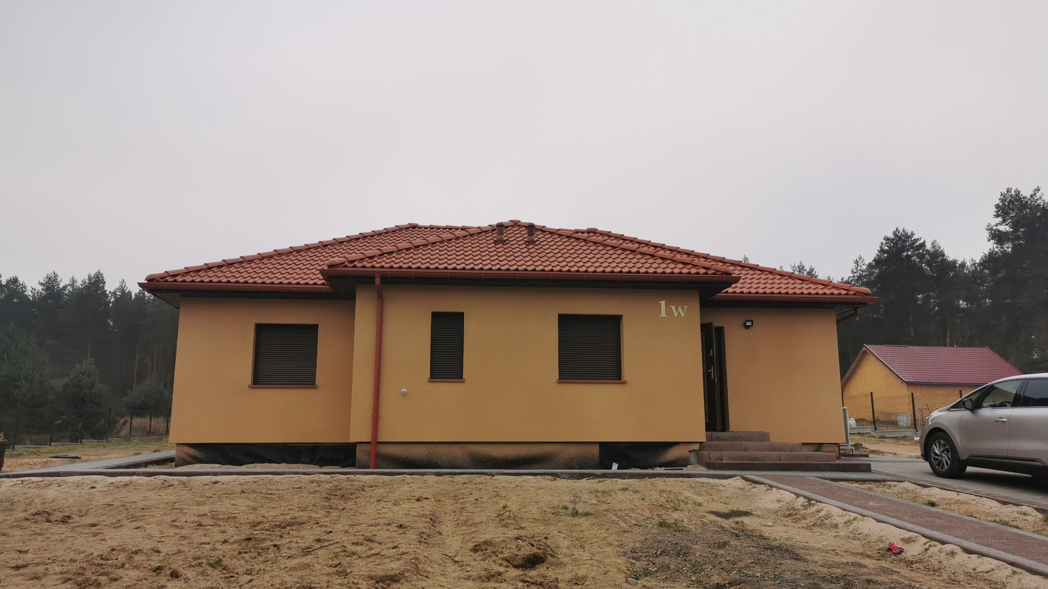 Parterowy dom murowany z brązową dachówką i numerem '1w' na elewacji, widok z poziomu gruntu na tle lasu i pochmurnego nieba.