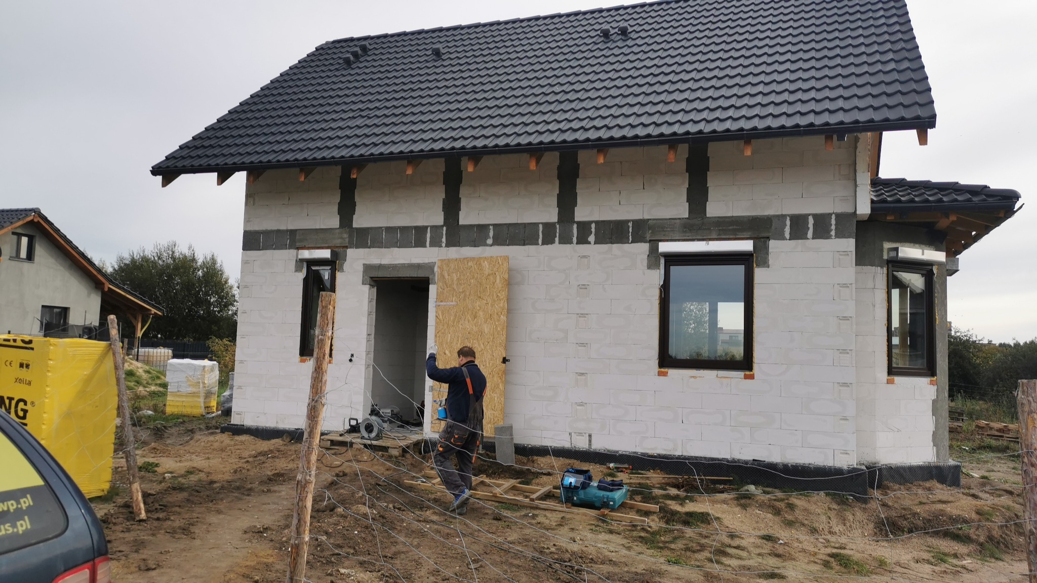 Budowa domu z białych bloczków, widoczny pracownik montujący tymczasowe drzwi z płyty OSB, czarny dach z dachówki, narzędzia budowlane na placu budowy.
