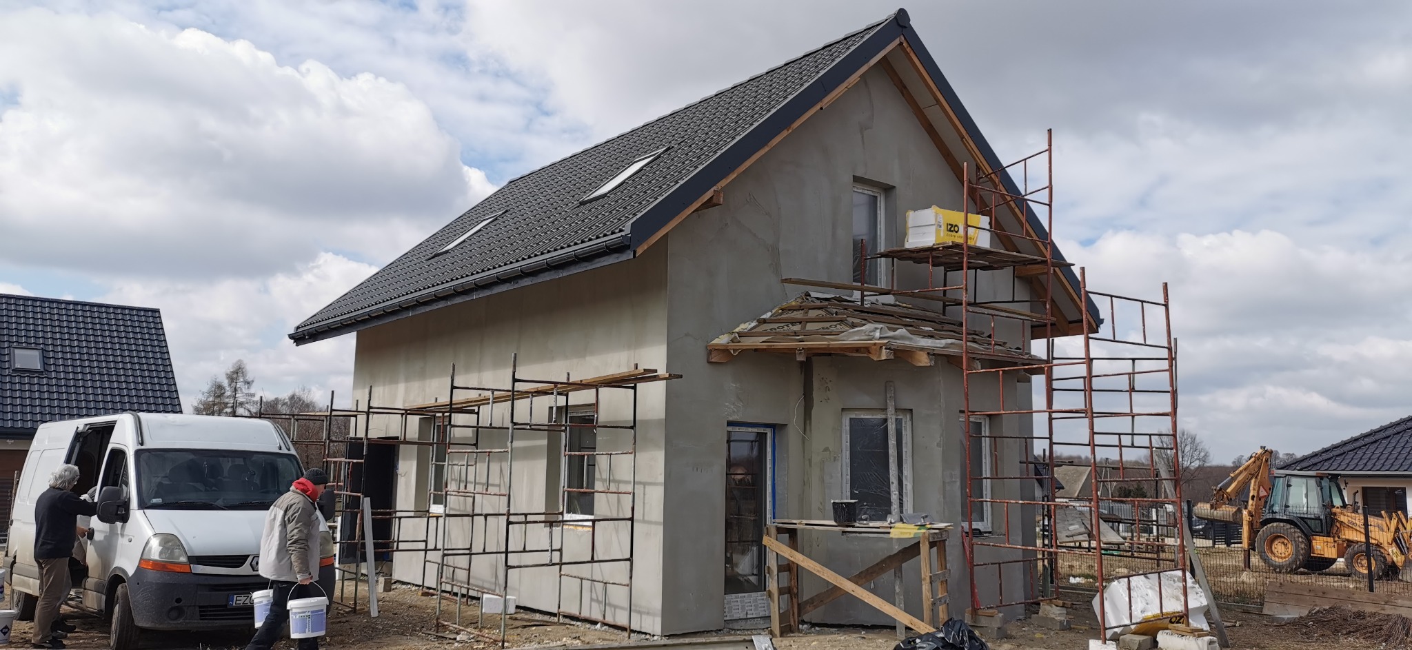 Surowy dom w trakcie tynkowania, rusztowania wokół budynku, ekipa budowlana przy białym busie, w tle koparko-ładowarka i inne domy.