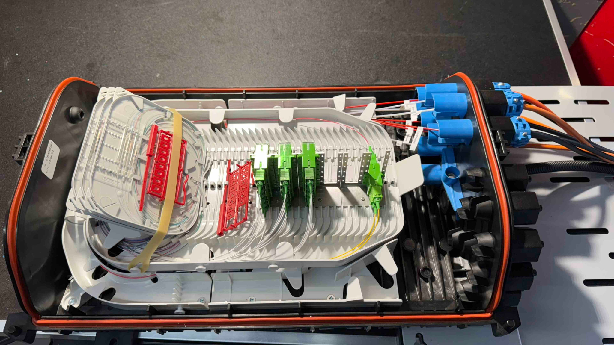 Otwarta skrzynka telekomunikacyjna z widocznymi światłowodami, złączami SC w kolorach zielonym i czerwonym oraz niebieskimi złączkami, ułożonymi wewnątrz białej kasety światłowodowej z pomarańczową...