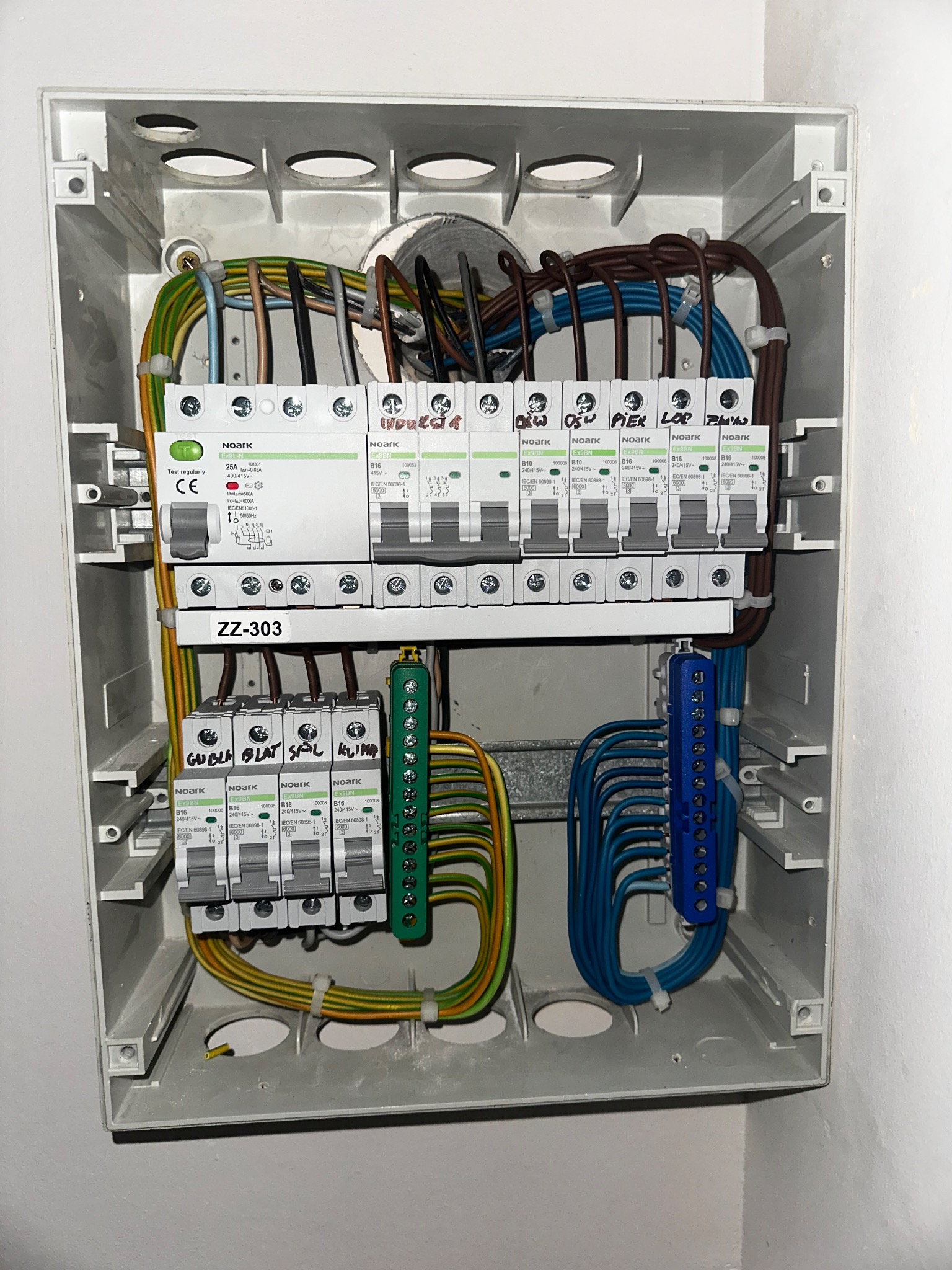 Otwarta skrzynka elektryczna z zainstalowanymi bezpiecznikami, okablowaniem i listwami zaciskowymi. Widoczne przewody w różnych kolorach i oznaczenia na elementach.