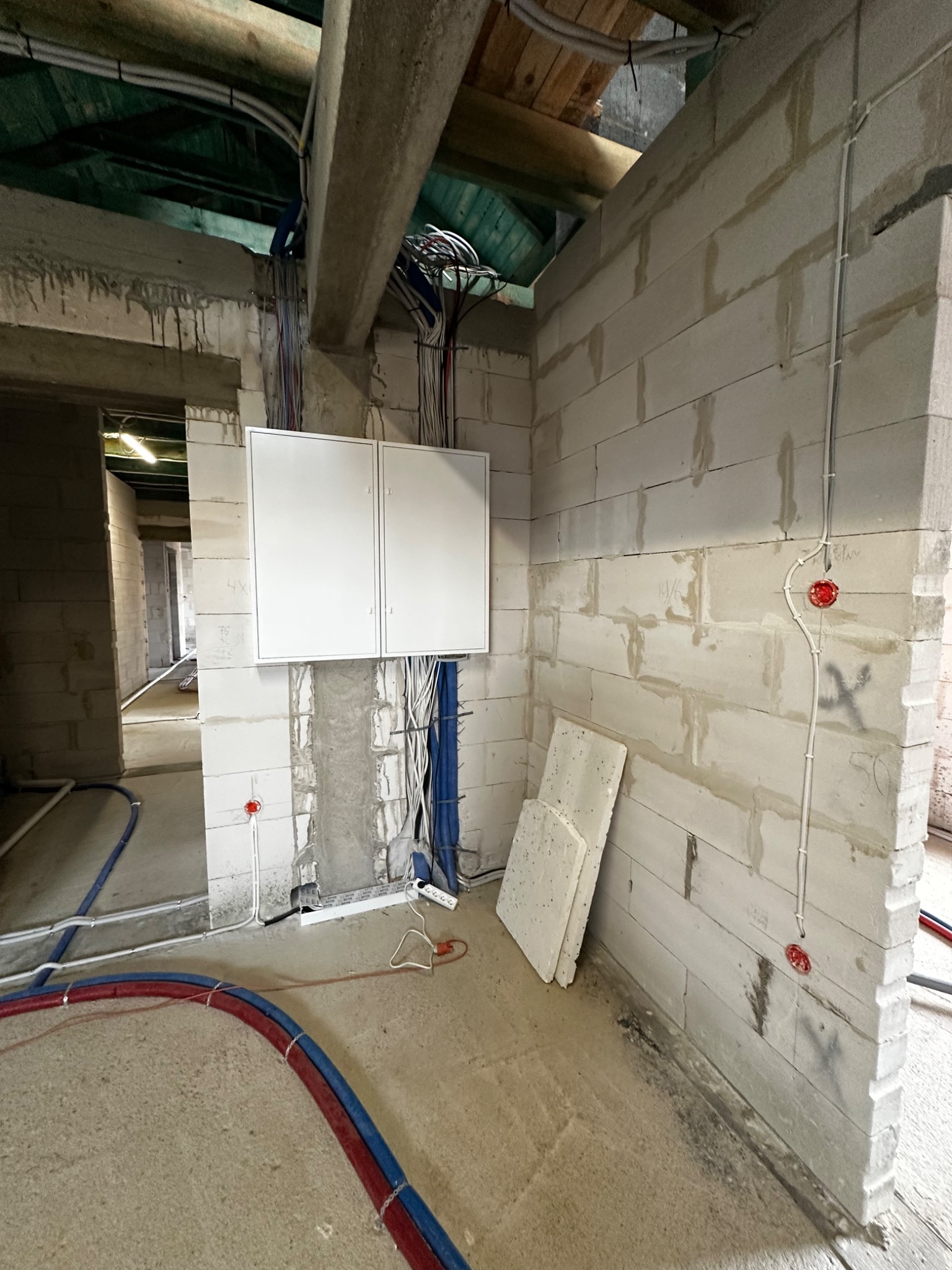 Instalacja elektryczna w surowym wnętrzu: biała skrzynka rozdzielcza, kable, puszki podtynkowe w ścianie z betonu komórkowego. Widoczne rury instalacji wodnej.