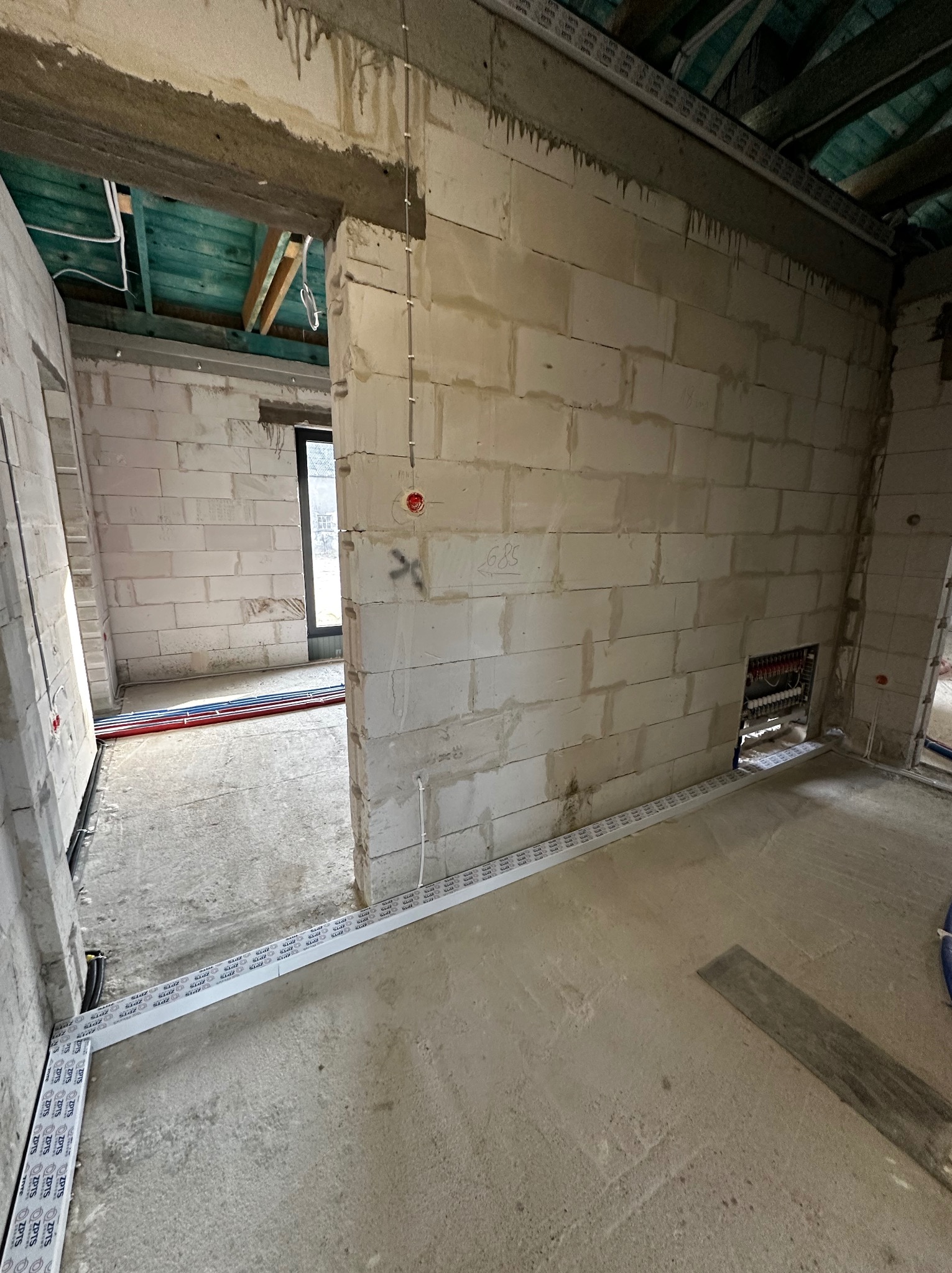 Surowy stan wnętrza z instalacjami: przewody elektryczne, rury grzewcze w ścianach z bloczków. Widoczne kable i peszle. Podłoże betonowe, surowy strop.