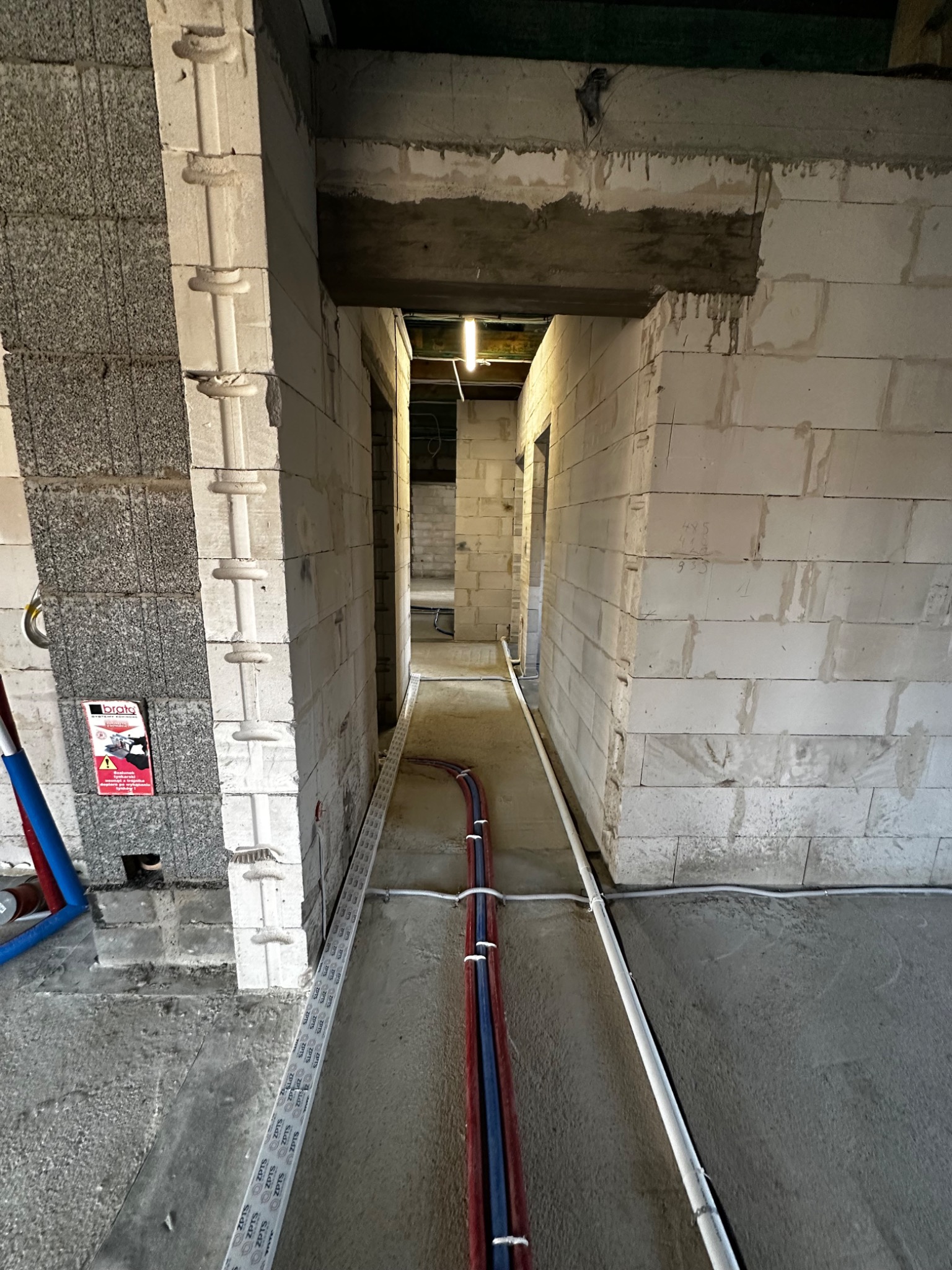Surowy stan wnętrza budynku w trakcie budowy. Widoczne ściany z betonu komórkowego, instalacje elektryczne i rury prowadzone wzdłuż podłogi. W tle widoczne kolejne pomieszczenia.
