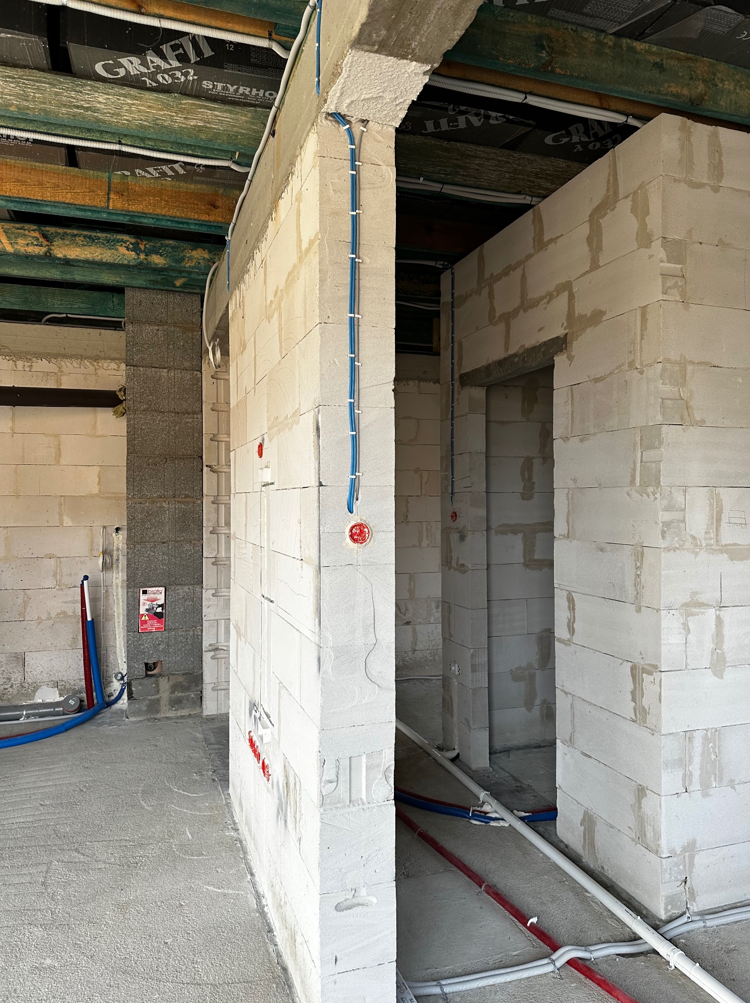Wnętrze w trakcie budowy z widocznymi instalacjami elektrycznymi w peszlach na ścianach z betonu komórkowego. Widoczne również rury instalacji wodnej w podłodze.