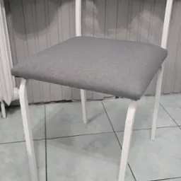 Krzesło z białym metalowym stelażem i szarym tapicerowanym siedziskiem na tle szarej ściany i zasłony.