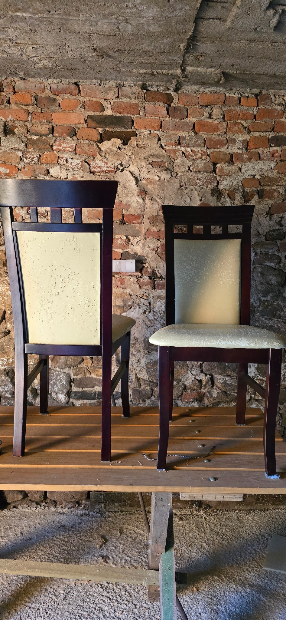 Dwa krzesła z ciemnego drewna z jasną tapicerką, stojące na drewnianej platformie na tle ściany z czerwonej cegły i kamienia.