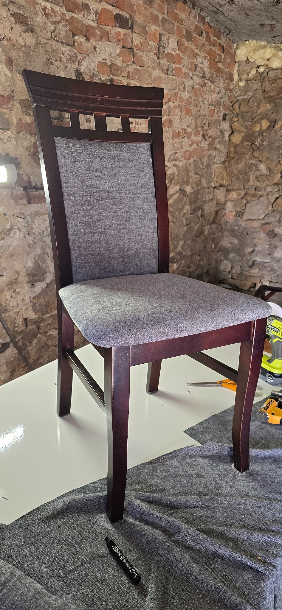 Krzesło z ciemnego drewna z nową, szarą tapicerką na siedzisku i oparciu, leżące na stole z narzędziami tapicerskimi i kawałkiem materiału w tle ściana z cegły