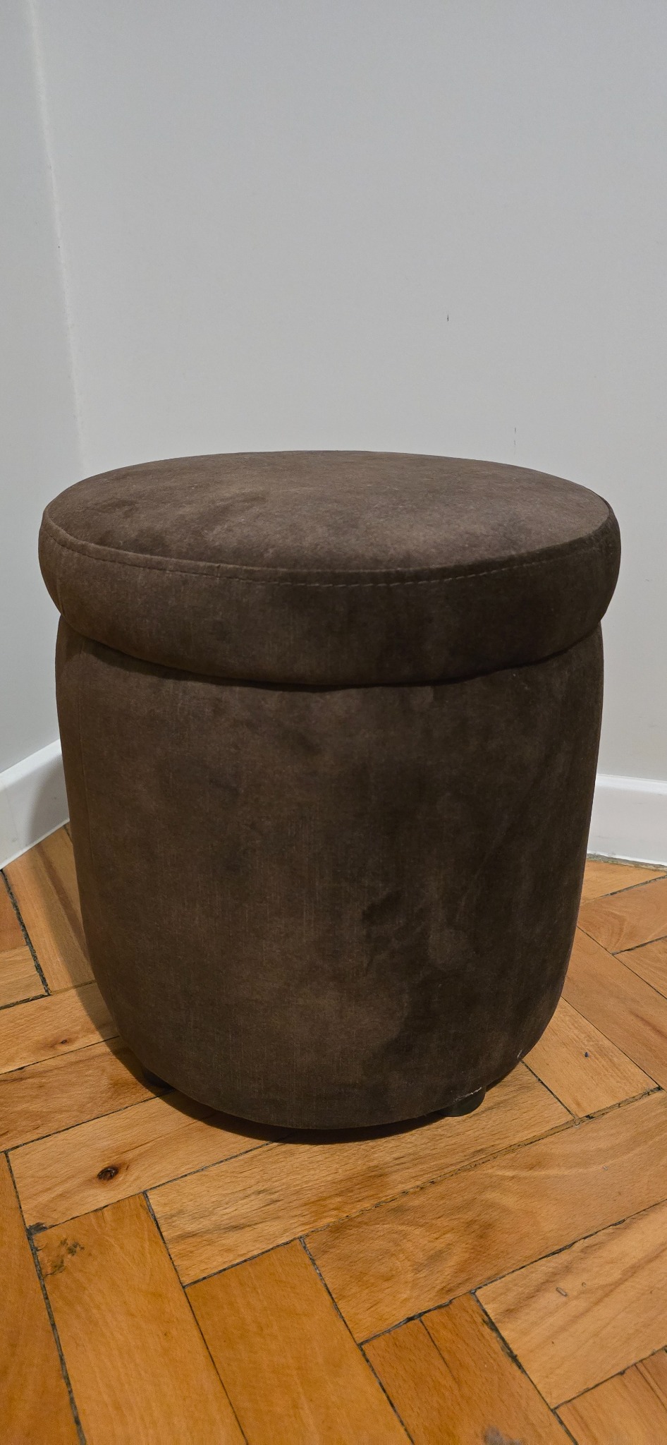 Brązowy, okrągły puf obity zamszem, stojący na parkiecie ułożonym w jodełkę, na tle białej ściany.