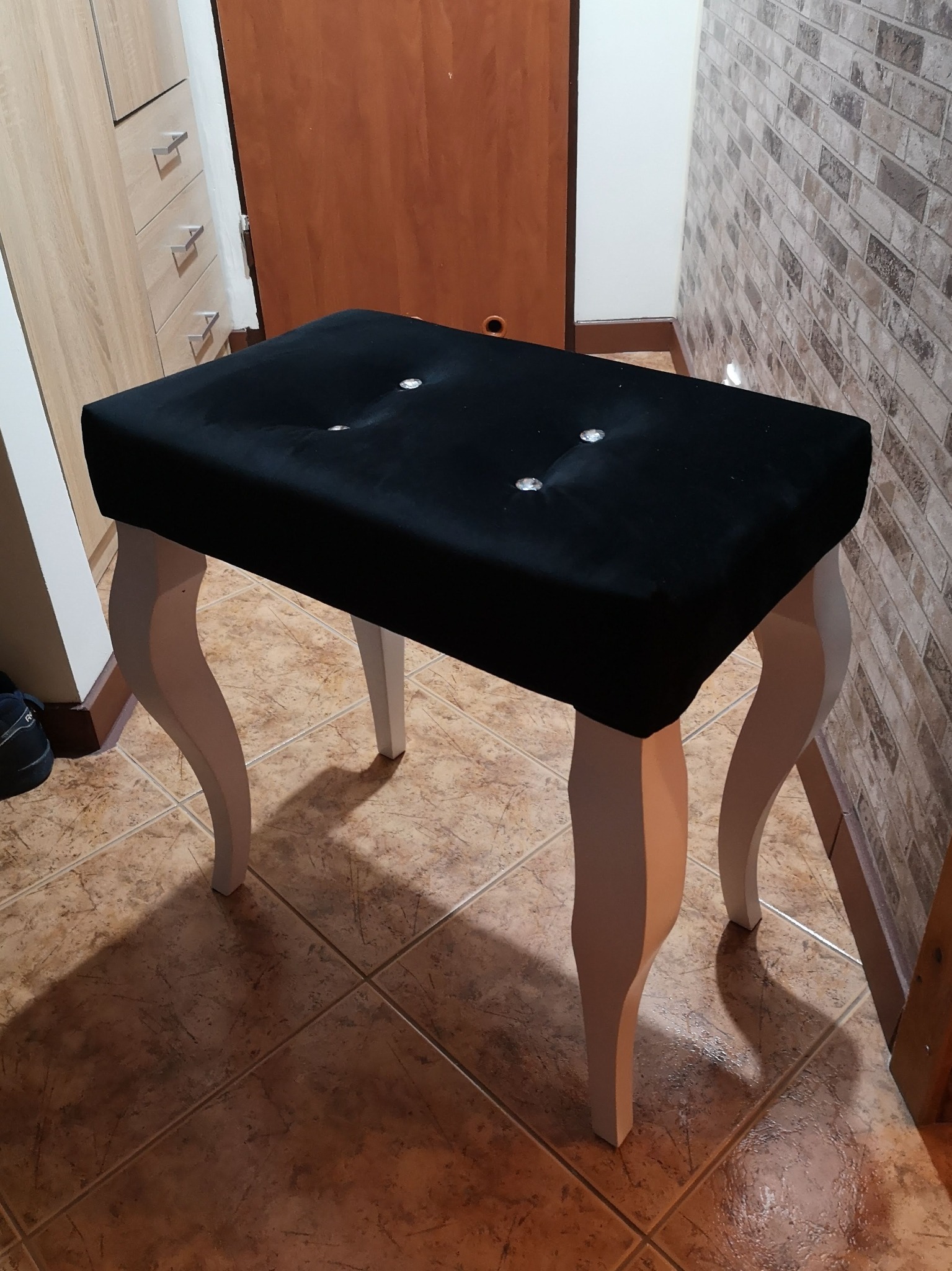 Tapicerowany taboret z czarnym siedziskiem z guzikami i białymi, giętymi nogami, ustawiony na brązowej podłodze z płytek.