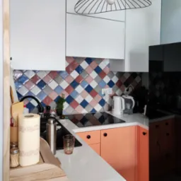Nowoczesna kuchnia z białymi górnymi szafkami, koralowymi dolnymi szafkami, blatem z białego lastryko i mozaiką z kolorowych płytek w romby na ścianie, oświetlona lampą wiszącą w kształcie klatki.