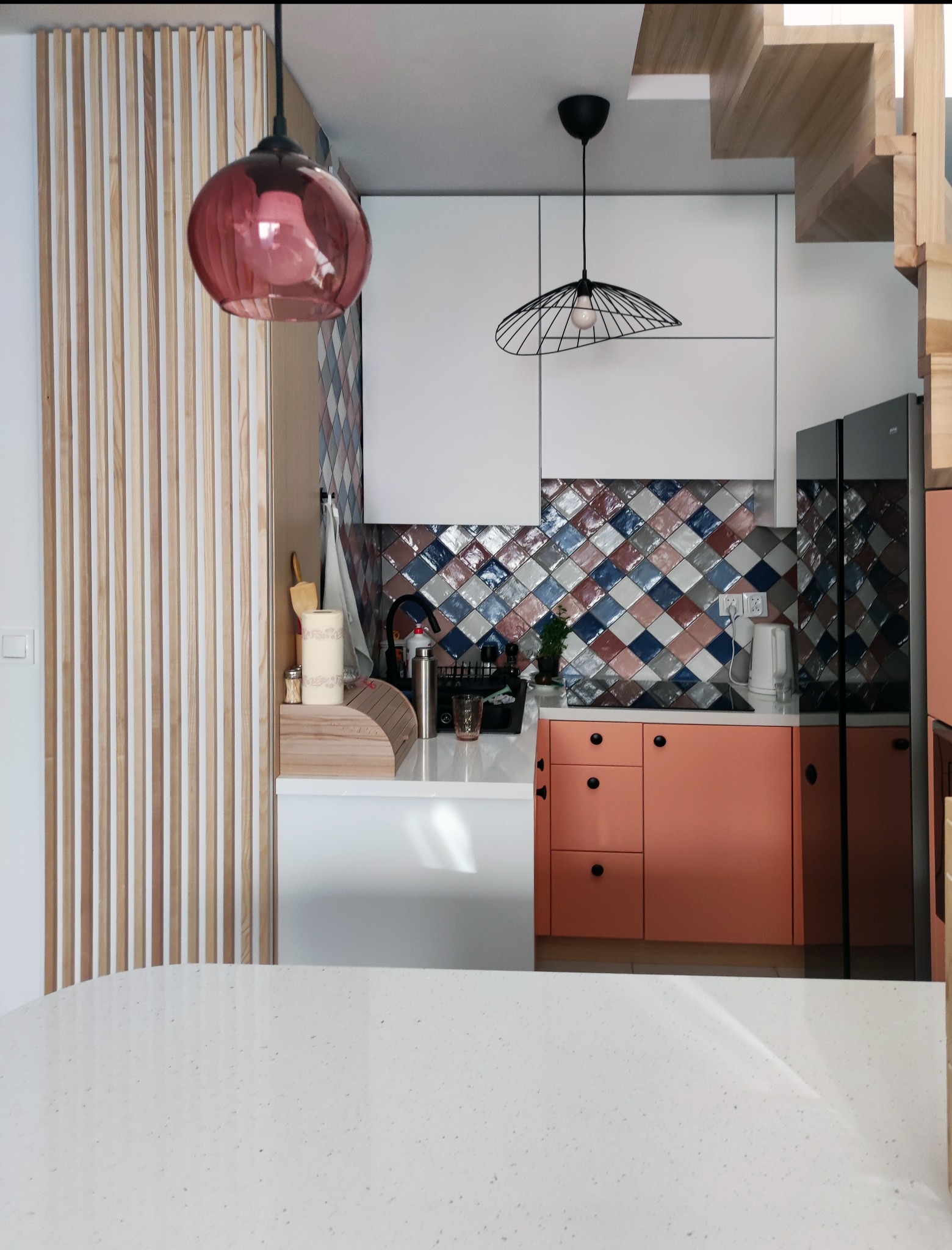 Nowoczesna kuchnia z elementami drewna, różowymi akcentami i wzorzystymi płytkami na ścianie, oświetlona wiszącymi lampami.
