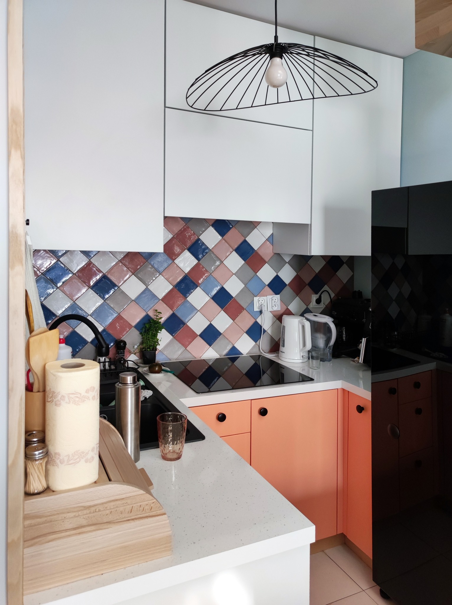 Nowoczesna kuchnia z białymi górnymi szafkami, koralowymi dolnymi szafkami, blatem z białego lastryko i mozaiką z kolorowych płytek w romby na ścianie, oświetlona lampą wiszącą w kształcie klatki.