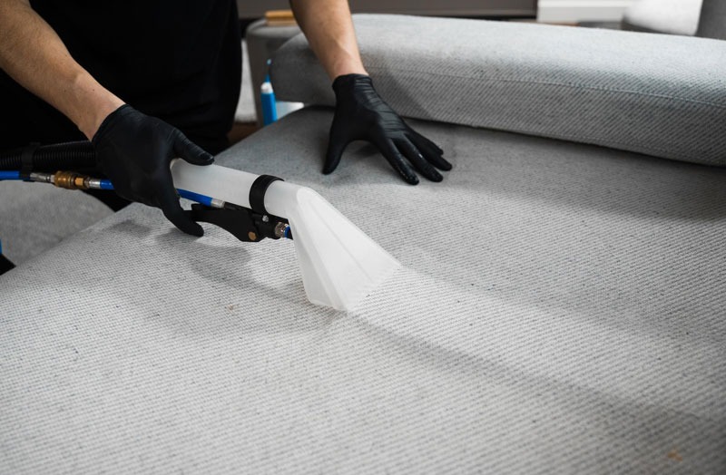 Czyszczenie jasnej sofy tekstylną dyszą parową przez osobę w czarnych rękawicach. Widoczny efekt czyszczenia na materiale.