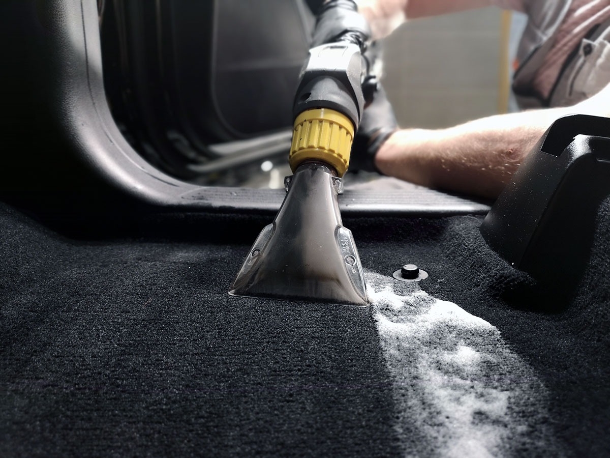 Czyszczenie tapicerki samochodowej: dysza odkurzacza ekstrakcyjnego usuwa zabrudzenia z czarnej wykładziny podłogowej, pozostawiając smugę piany.