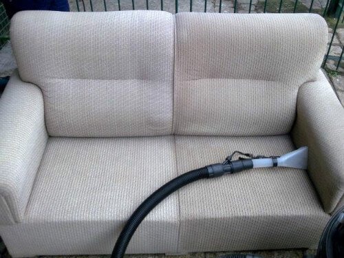 Jasna, dwuosobowa sofa z dyszą odkurzacza piorącego na siedzisku. Widoczna struktura materiału i czarny wąż odkurzacza. Ujęcie z bliska, naturalne światło.