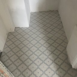 MASTERBUD MAKSYM ROZUMNIAK - Ułożona podłoga z szarych płytek o marokańskim wzorze w trakcie prac wykończeniowych w małej łazience, widoczne niedociągnięcia przy ścianach i niezagruntowane ściany.