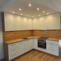 MASTERBUD MAKSYM ROZUMNIAK - Nowoczesna, narożna kuchnia z białymi szafkami i drewnianym blatem oraz panelem ściennym, oświetlona punktowymi lampami sufitowymi, z widocznym zlewem i piekarnikiem.