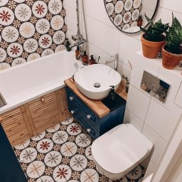 MASTERBUD MAKSYM ROZUMNIAK - Wnętrze wykończonej łazienki w Warszawie z wanną obudowaną drewnianymi lamelami, umywalką nablatową na niebieskiej szafce z drewnianym blatem, toaletą oraz wzorzystymi płytkami podłogowymi...